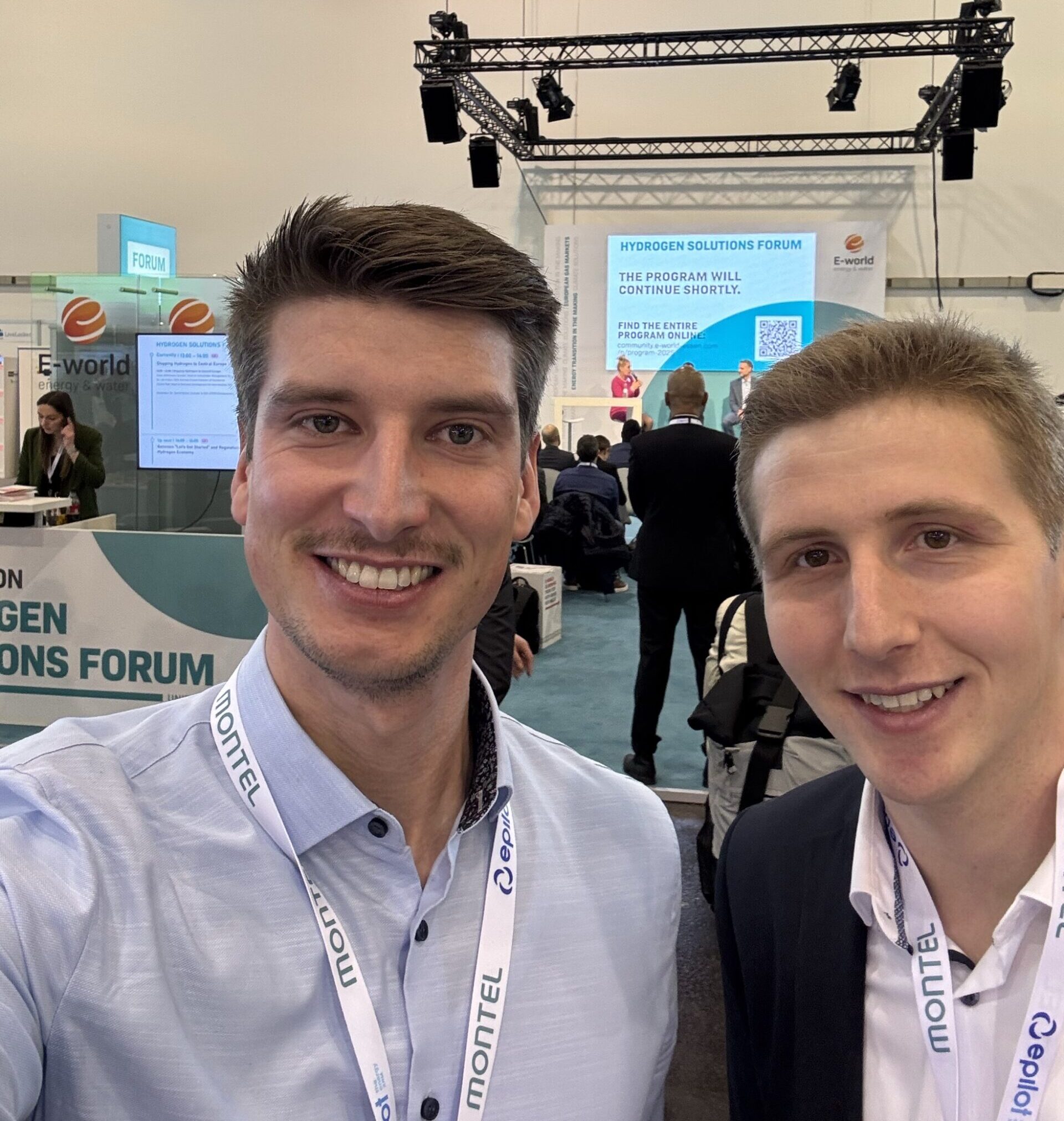 Zwei Männer lächeln in die Kamera, im Hintergrund eine Ausstellung mit Bildschirmen