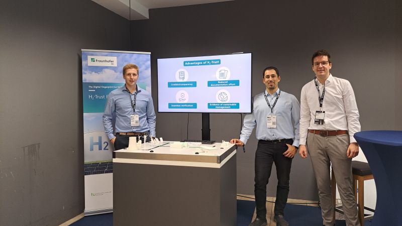 drei Männer vor einem Blockchain-Wasserstoff-Demonstrator, Bildschirm und Roll-up im Hintergrund