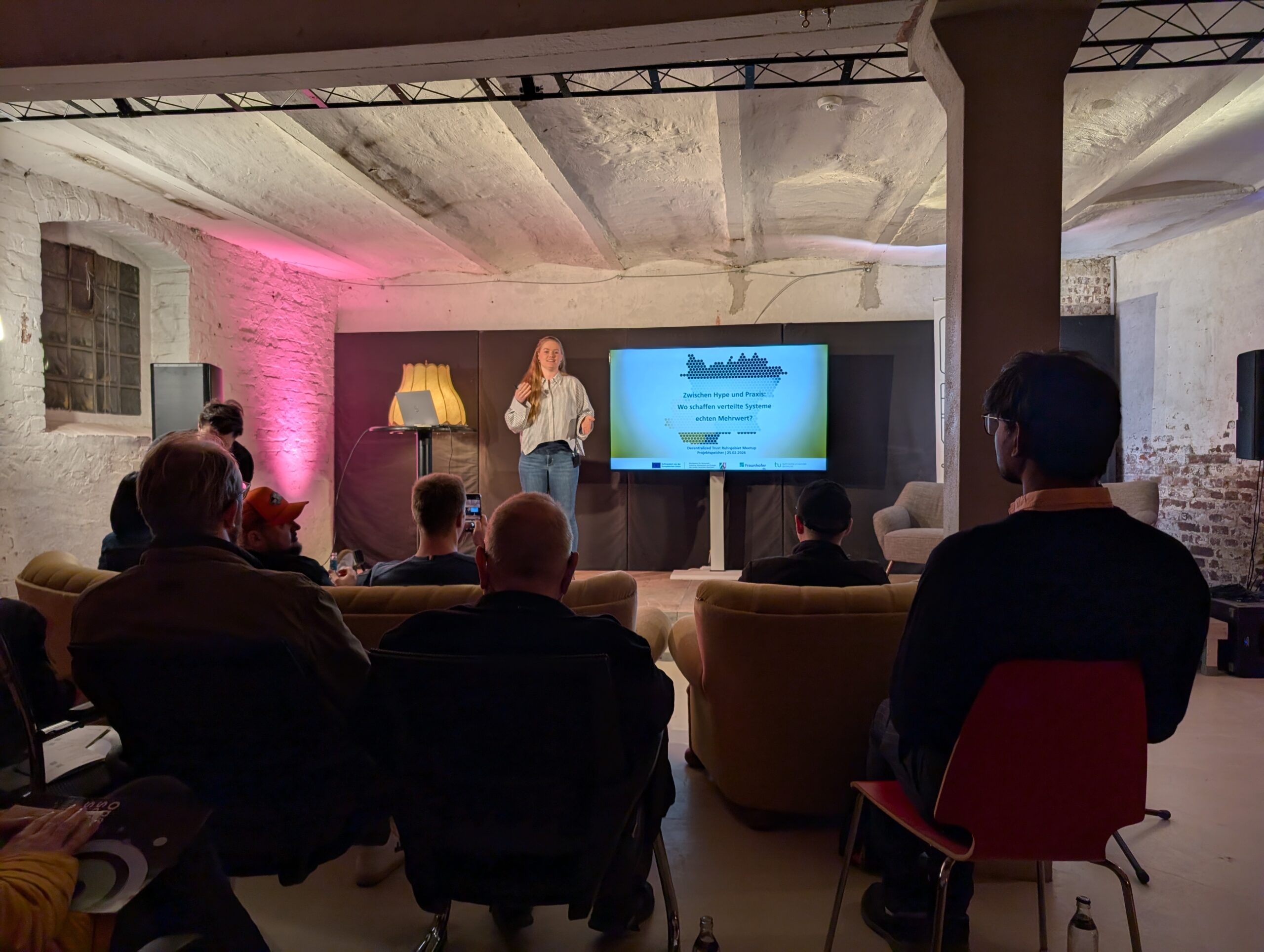 blonde Frau steht auf einer Bühne in einer Industriehallte, Publikum sitzt auf Stühlen und Couches davor, Bildschirm zeigt Präsentation.